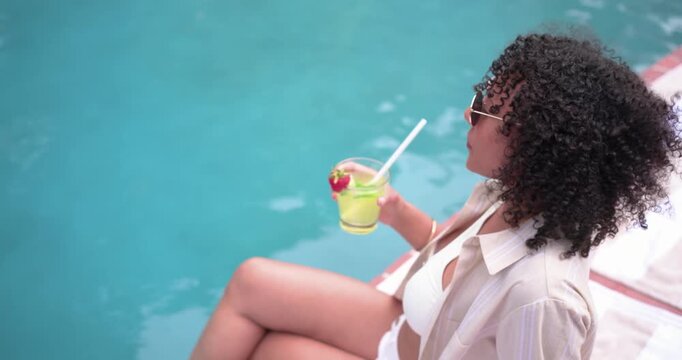 Woman is tapping straw, lifting green cocktail glass and sipping while posing and smiling poolside