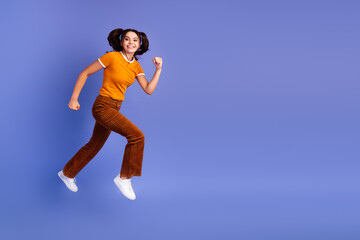 Fototapeta premium Young girl jumping in the air smiling orange shirt brown pants white sneakers against violet background