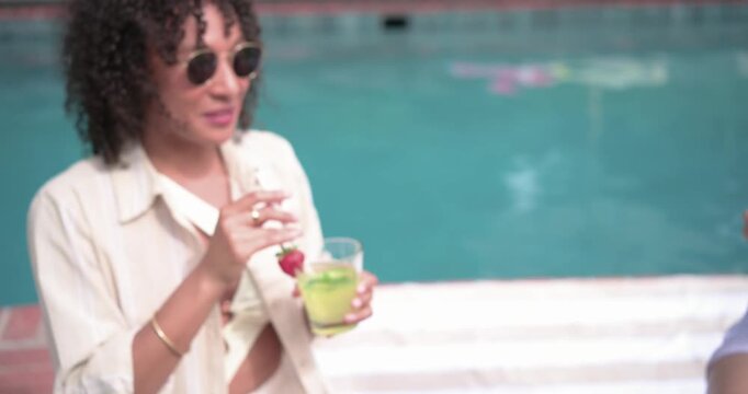 Diverse women friends clinking green drinks with straws, adjusting sunhat and chatting poolside