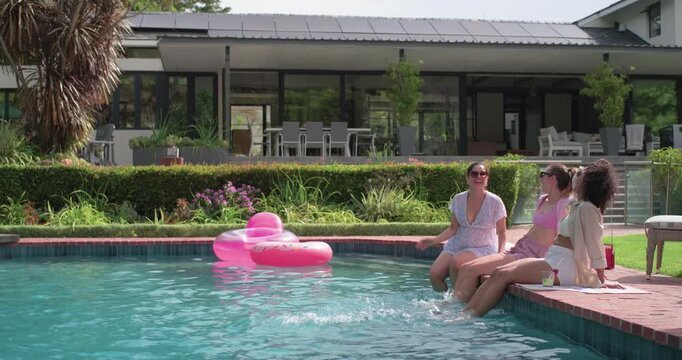 Diverse female friends arriving sitting on brick coping pool, dipping feet cooling off, copy space