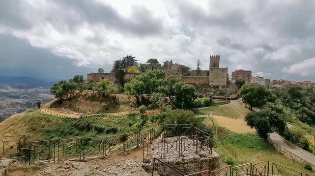 Scenic view of Lombardy Castle in Enna, Sicily, showcasing the castle's architecture, surrounding greenery, and distant hills under a cloudy sky