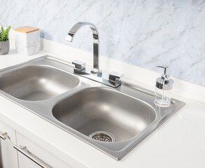 Fototapeta premium Close-Up of Double Basin Drop-In Stainless Steel Kitchen Sink and Faucet Mounted on a White Sink Base Cabinet in Bright Modern Kitchen, White Marble Background.