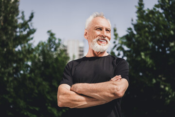 Friendly senior man in black athletic shirt stands outdoors with arms crossed smiling confidently in a park setting near the city skyline © deagreez