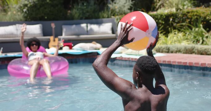 African American friends playing home pool, man raising tossing beach ball to woman on pink ring