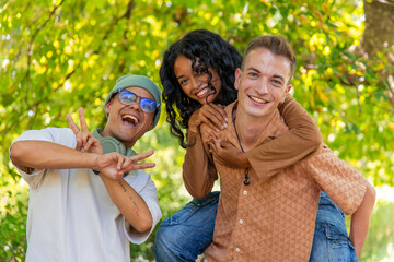 Fototapeta premium Three cheerful multi-ethnic friends are enjoying a sunny day in the park. With two of them giving a piggyback ride and the other one gesturing peace signs. Symbolizing their joyful and carefree mood