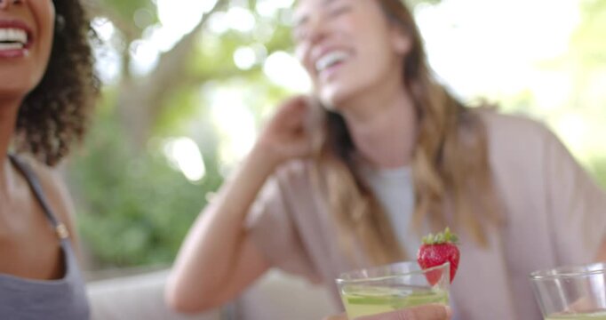 Diverse female friends clinking glasses with lime and strawberry, celebrating on shaded patio