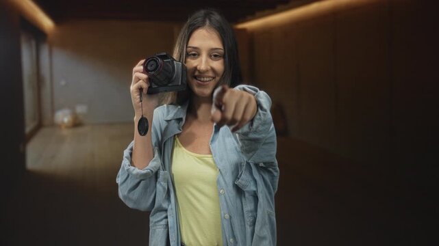 Young hispanic woman smiling and holding camera with lens cap dangling, pointing finger to lens in studio setting; playful connection.