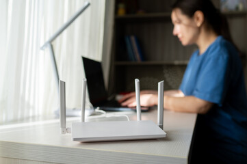 White WiFi Router in Foreground with Woman Working on Laptop in Blurred Background at Home Office