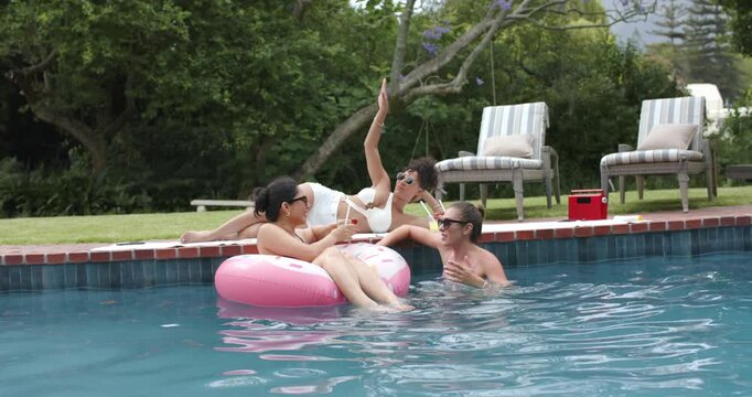 Three women in swimwear passing strawcups on pink float reaching after deck woman raising arm pool