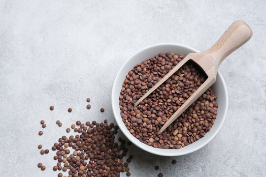 Brown lentils in a white bowl with a wooden scoop, scattered lentils on a gray surface, showcasing natural food elements and textures for culinary use