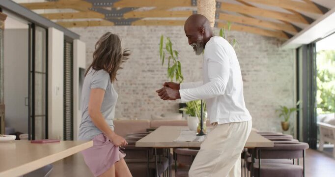 Diverse couple playing in open kitchen after man raising knee, tugging shorts by dining table vase