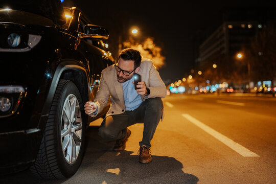 Man examining a flat tire on his car at night, using his phone's flashlight for illumination on an urban street