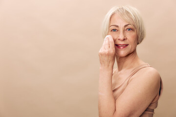 Mature woman with short blonde hair gently cleansing her face with a cotton pad on beige background. Concept for skincare, beauty routine, anti aging and self care.