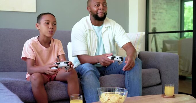 African American father and child high-fiving, grabbing chips, playing on white controllers at home