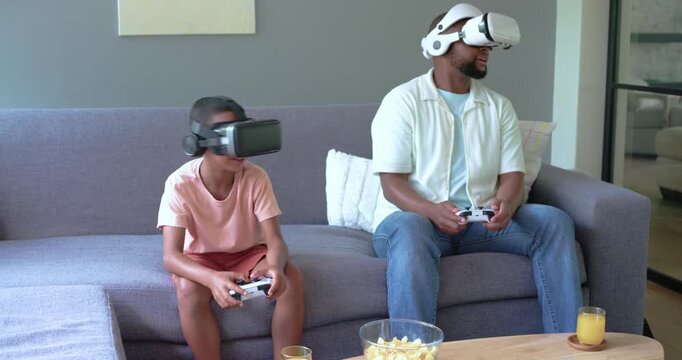 African American father and teen son playing VR game, pressing controller for fun on sofa headsets