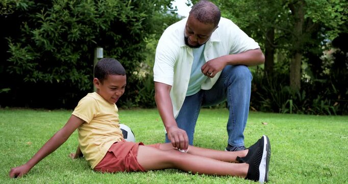 African American father kneeling, dabbing and bandaging school-age son's shin after scrape on lawn