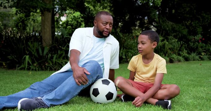 African American father and son on lawn son showing knee bandage father soothing near soccer ball
