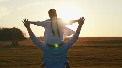 Dad daughter dream of flying together. Little daughter on father's shoulders play together in park, sun sky, silhouette. Family outdoor activity. Happy family. Dad, kid daughter walk in nature, sunset © Victoriia
