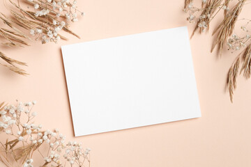 Blank invitation or greeting card mockup on a soft peach background surrounded by dried flowers, creating a minimalist and elegant presentation for design purposes