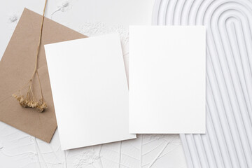 Wedding invitation card mockup with two blank white cards and craft brown envelope, placed on textured white background with dried flower decor and curved lines