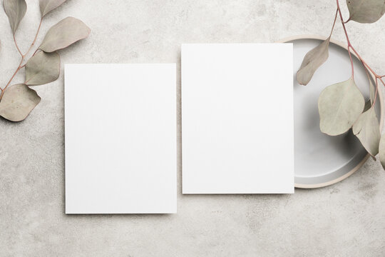 Wedding invitation card mockup featuring two blank cards on a textured surface with natural dry eucalyptus leaves and a round plate in a minimalist setting