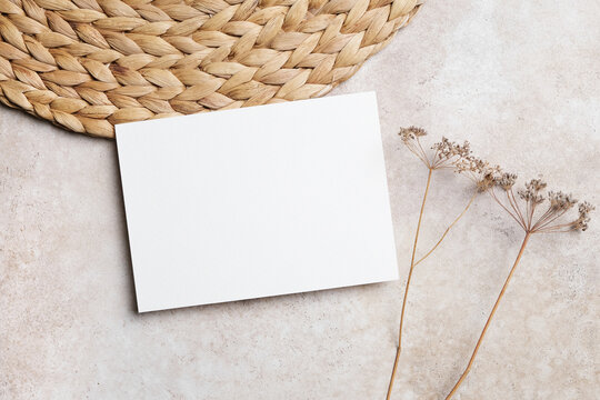 Blank invitation or greeting card mockup stony background with botanical decor and natural straw placemat. Boho style flat lay with copy space for card decor