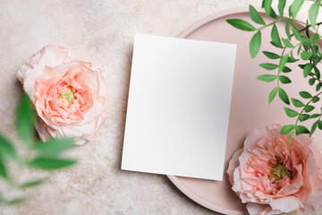 Vertical white blank card on a round beige plate with peach roses and lush green leaves. Elegant flat lay, perfect for menu, invitation, or greeting card designs