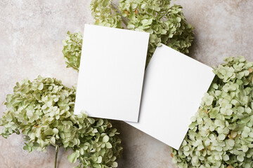 Two blank white greeting cards mockup placed among green hydrangea flowers on a neutral stone background, perfect for showcasing elegant stationery designs