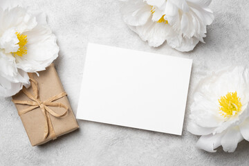Blank greeting card mockup with copy space surrounded by white peonies and a wrapped gift on a textured gray surface for creative design projects