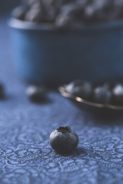 Single Blueberry with Spoon and Bowl Full of Blueberries out of Focus in Background on Blue Fabric