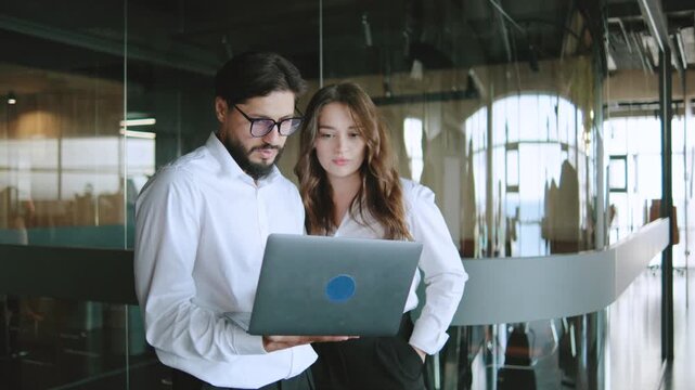 Colleagues review project details on a laptop in a modern office environment during working hours with large windows and natural light