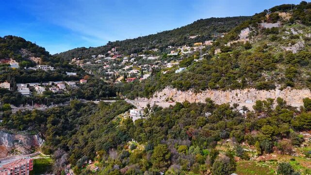 Road on the mountain slope is crossed by the cars. Slopes of the green rocks with the cityscape of Nice, France. Aerial view.