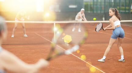 Back view of concentrated young female tennis player engrossed in game on clay court, swinging racket ready to hit ball over net during doubles with female partner and opponents captured in background © JackF