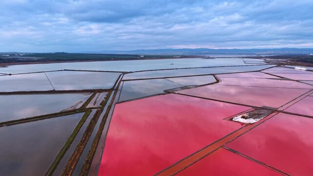 Large pink salt evaporation pond in coastal landscape. Aerial perspective of bright pink saltern pond against blue water and distant mountains.