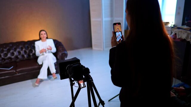 Over the shoulder view of smartphone filming interview. Person recording woman in white suit using mobile phone in professional studio with camera.