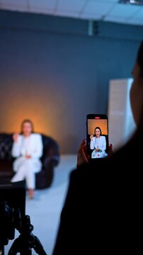 Behind the scenes of video recording on smartphone. Person holding smartphone recording woman in white suit sitting on leather sofa in professional studio environment.