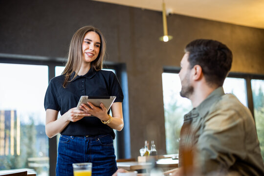 Smiling waitress taking a digital order from a young man sitting at a restaurant table.
