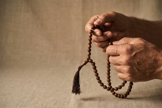 Elderly man's hands holding and counting wooden prayer beads (tasbih/misbaha) against a linen background.