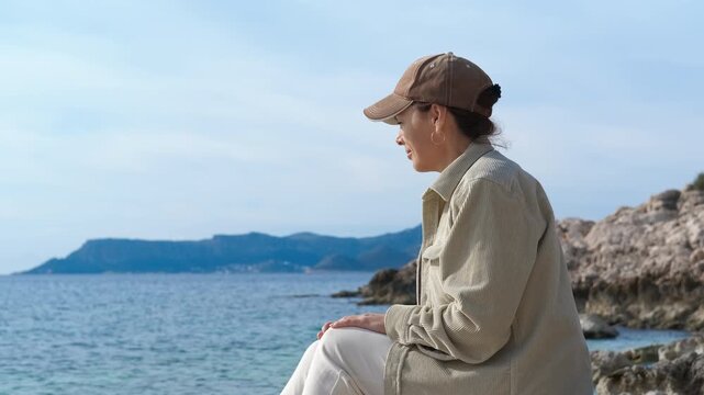 Tourist enjoying scenic ocean view from rocky coast. Female tourist wearing casual clothes and baseball cap sits on rocky coast, enjoying serene ocean view with distant islands under clear sky