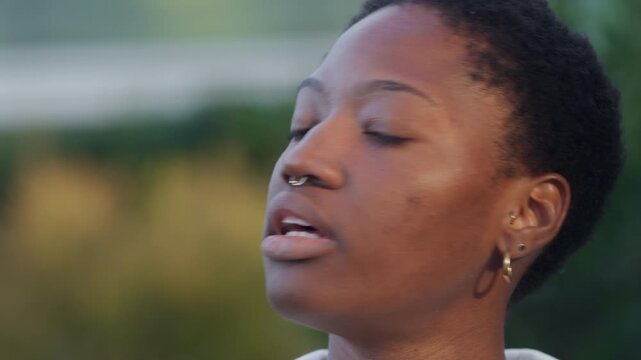 Closeup shot of black woman smoking cigarette outdoors. Young female exhaling smoke while looking at camera against blurred background