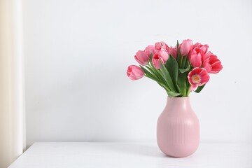 Obraz premium Bouquet of pink tulips in vase on table near white wall. Space for text