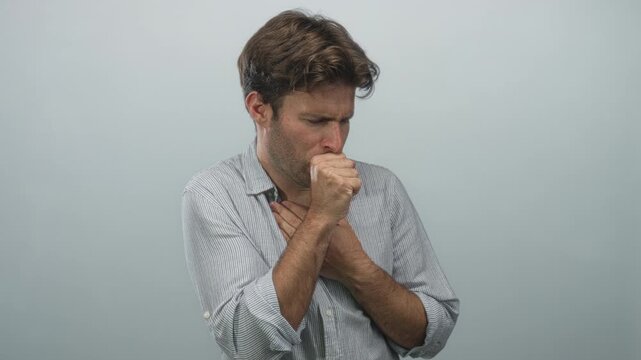 Young hispanic man with hand on chest and fist to mouth coughing in gray studio; illness discomfort.