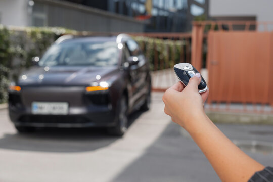 Woman stands holding a car key remote unlocking her parked car in an urban setting during daytime.