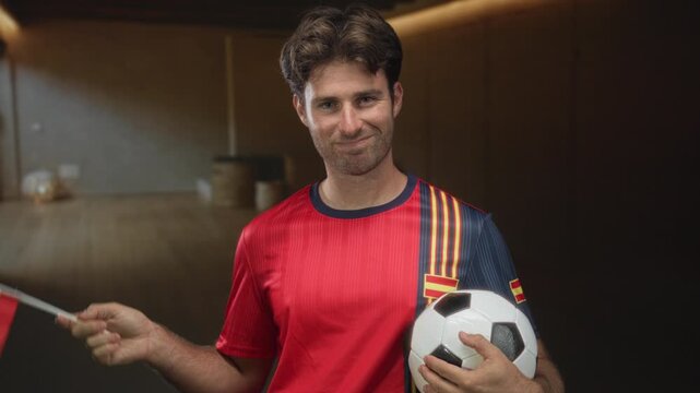 Young man holding a soccer ball and waving a spanish flag in studio while wearing a red and navy team jersey; national pride.
