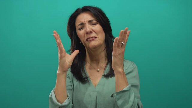 Middle aged woman raises hands to face in teal studio setting with expressive gesture and closed eyes showing despair; frustration.