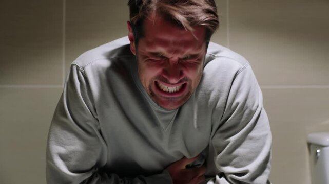 A Caucasian man sits on a toilet, grimacing intensely and clutching his stomach in discomfort, symbolizing severe abdominal pain or digestive distress.
