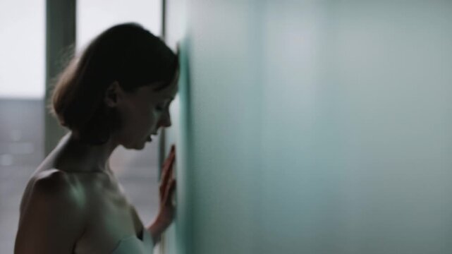 A young woman with short brown hair leans her head against a cool green wall, her hand touching the surface, conveying feelings of sadness and introspection.