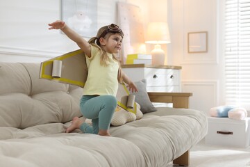 Cute little girl with cardboard plane wings and goggles playing pilot on sofa at home