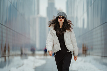 Woman in white puffer jacket walking in New York City. Front view of woman wearing white jacket and beanie walking along glass walls with city background © white78