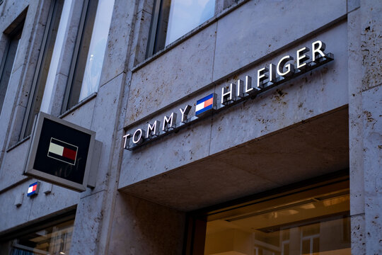 Luxury brand Tommy Hilfiger storefront with illuminated 3D lettering, iconic flag logo, mounted on textured beige stone facade, premium fashion retail presence in Germany, Hannover
